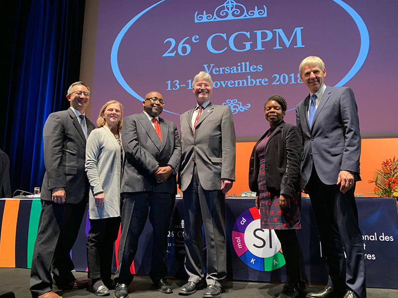 6 people standing on a stage in front of a screen that reads: 26th CGPM